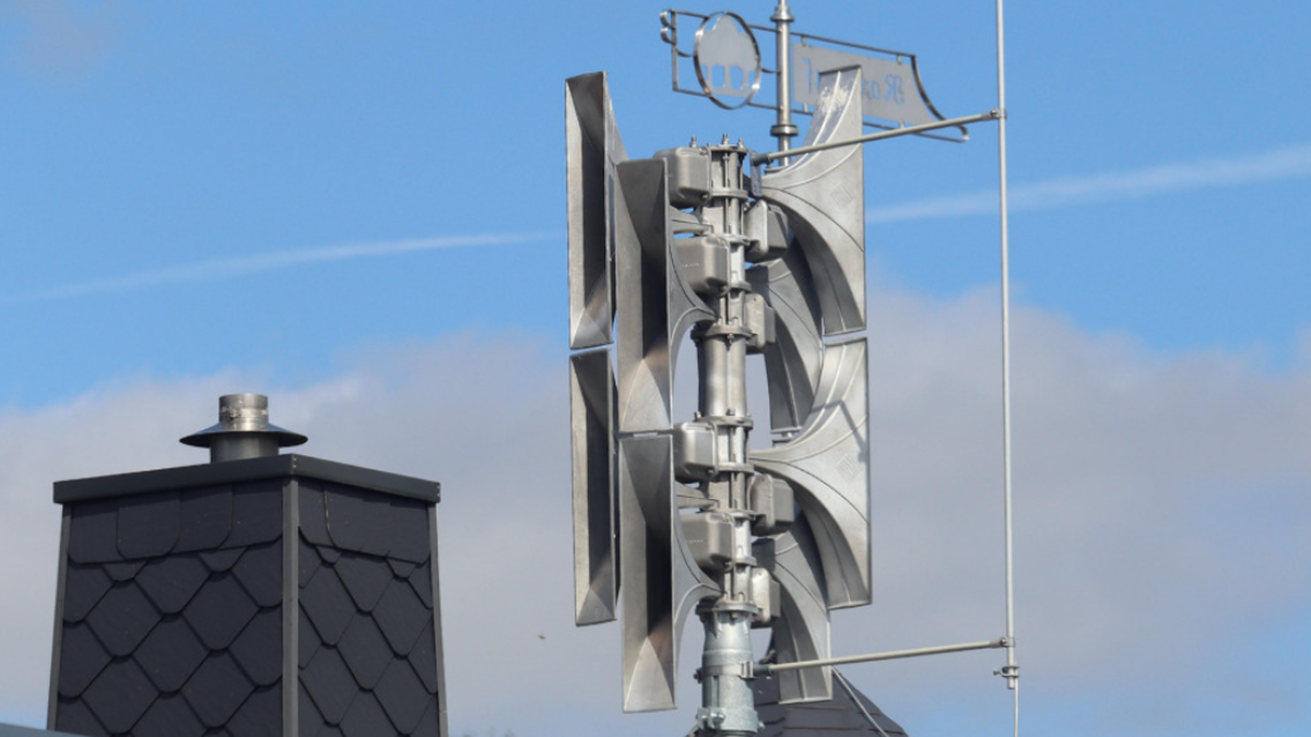 Wielostopniowa syrena alarmowa wykonana z błyszczącego metalu, zamontowana na maszcie na tle błękitnego nieba z niewielkimi chmurami. Obok syreny widoczny fragment dachu i komin budynku.
