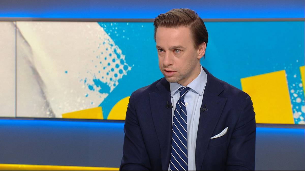 Mężczyzna w granatowym garniturze i pasiastym krawacie siedzi w studiu telewizyjnym.