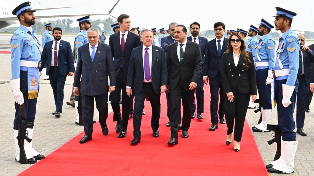 Grupa mężczyzn w garniturach idzie po czerwonym dywanie przed samolotem, wśr&oacute;d nich mężczyzna w niebieskim mundurze.