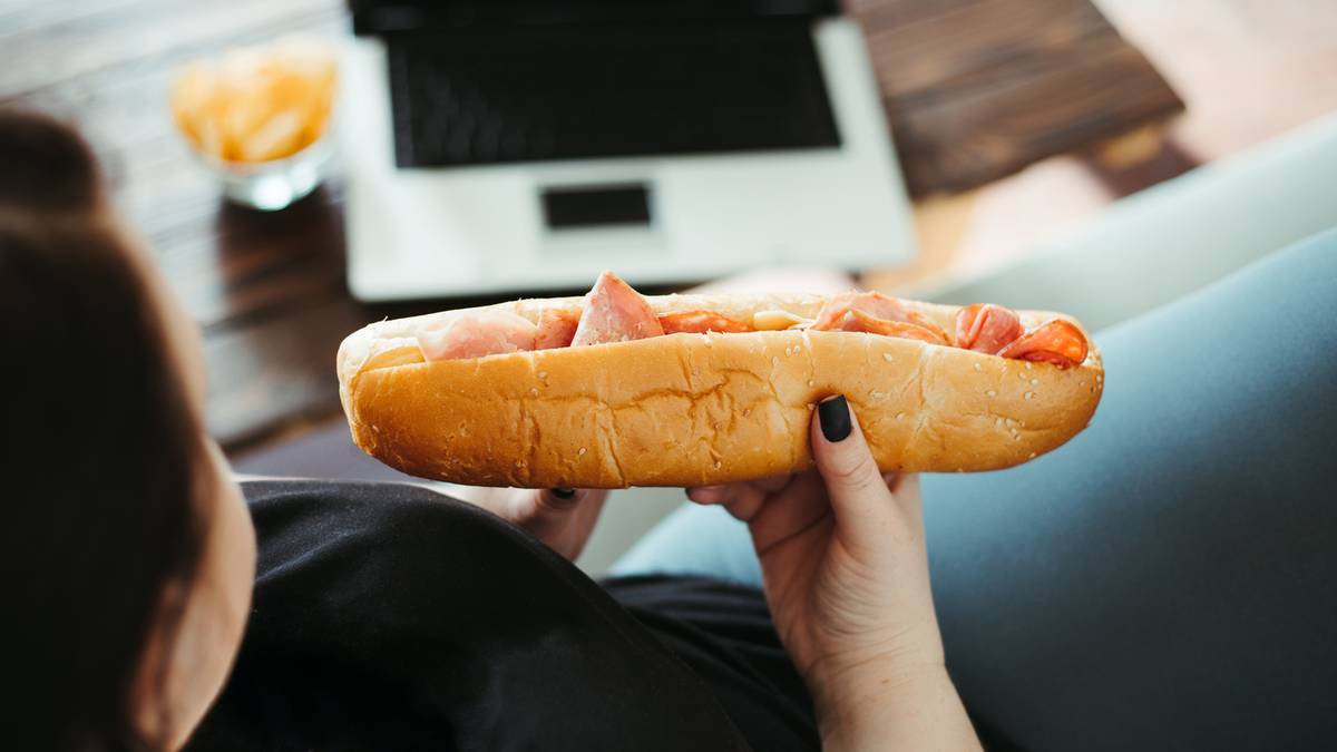 Osoba trzymająca par&oacute;wkę w bułce przed laptopem.
