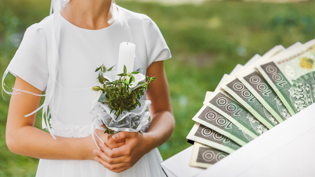 Dziecko w stroju komunijnym trzyma bukiecik ze świecą, obok stos banknot&oacute;w 100-złotowych.