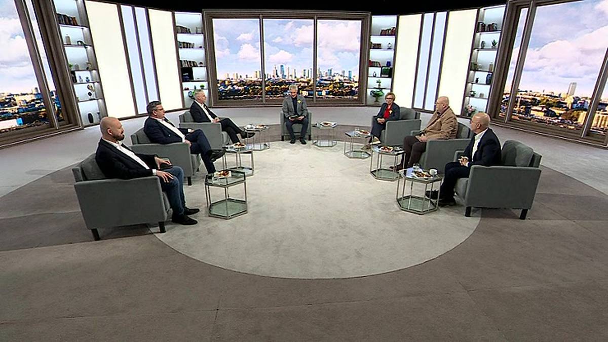 Siedem os&oacute;b w garniturach siedzi na kanapach w p&oacute;łokręgu, prowadząc dyskusję w studiu telewizyjnym. W tle widoczna panorama miasta.