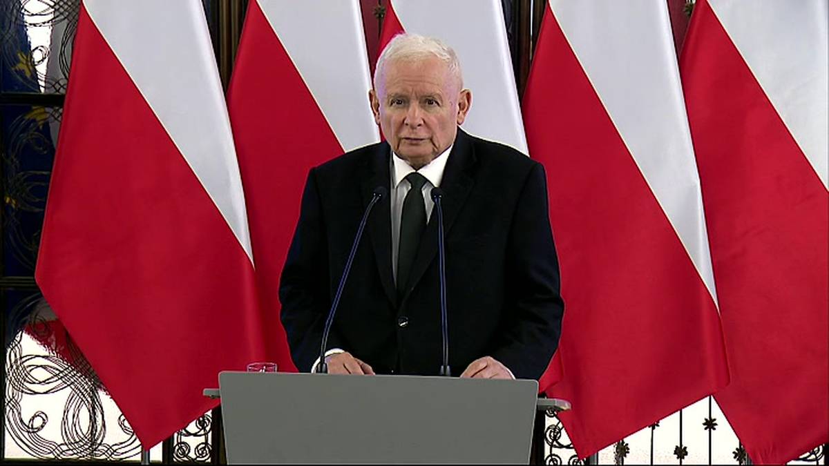 Jarosław Kaczyński przemawia za m&oacute;wnicą na tle polskich flag.