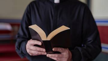 Duchowny w czarnej sutannie trzyma otwartą Biblię w dłoniach.