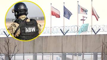 Funkcjonariusz ABW w kamizelce taktycznej z napisem "ABW", na tle budynku lotniska z flagami państwowymi