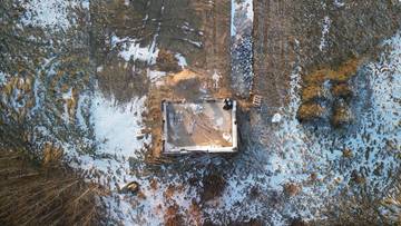 Plac budowy domu z widocznymi fundamentami i częściowo postawioną ścianą, przykrytymi plandeką, zimą.