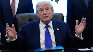 Donald Trump siedzi przy biurku, m&oacute;wiąc i gestykulując rękami.