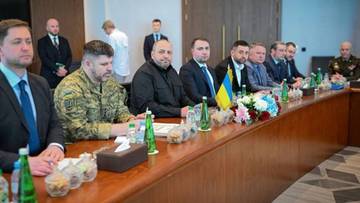 Grupa mężczyzn siedzi przy stole podczas spotkania delegacji, na stole ukraińska flaga i butelki z wodą.